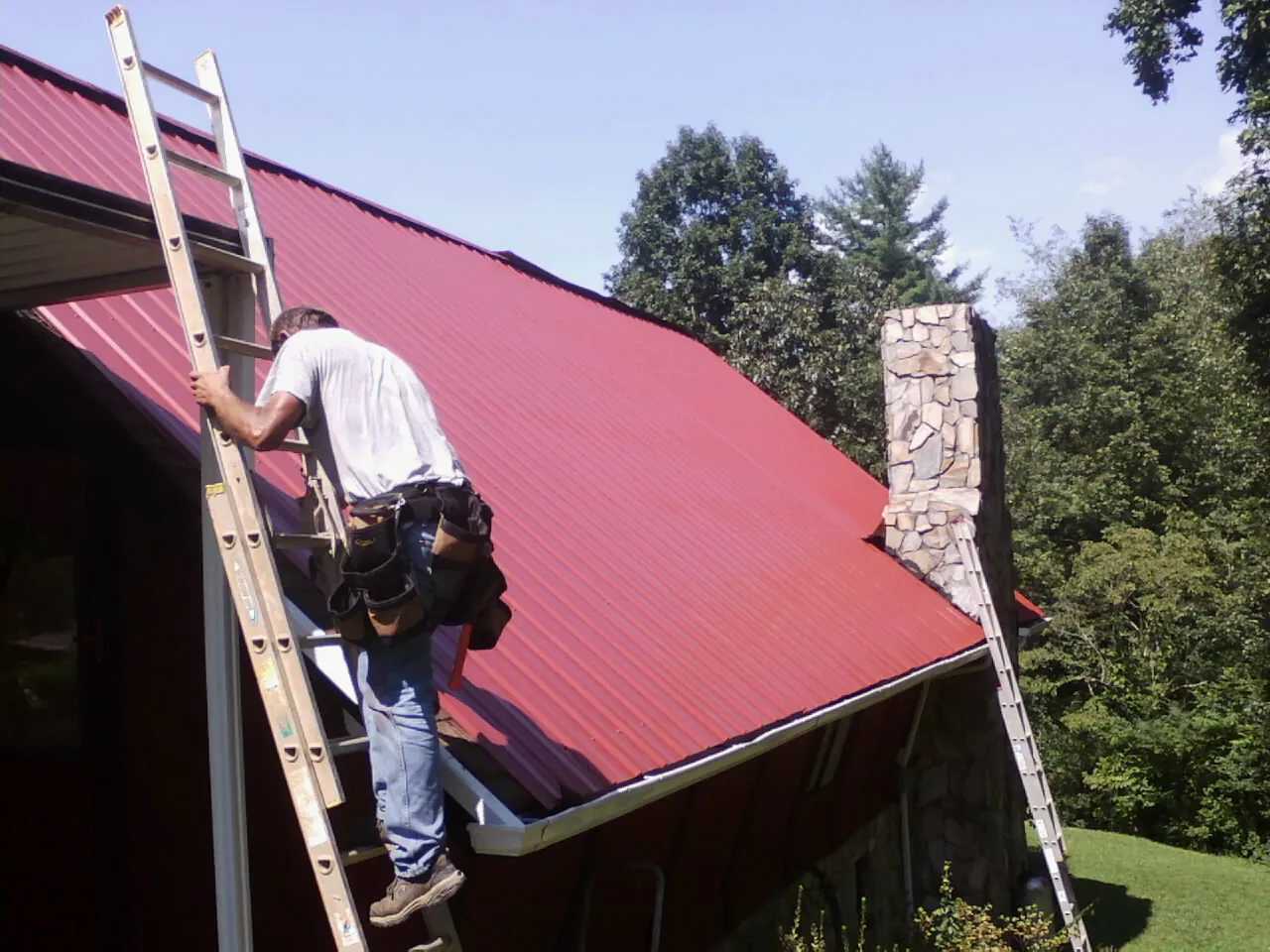 Roofer on a ladder beside a red metal roof and stone chimney — Soffit & Fascia Repair by Roofing Brothers in Petersburg